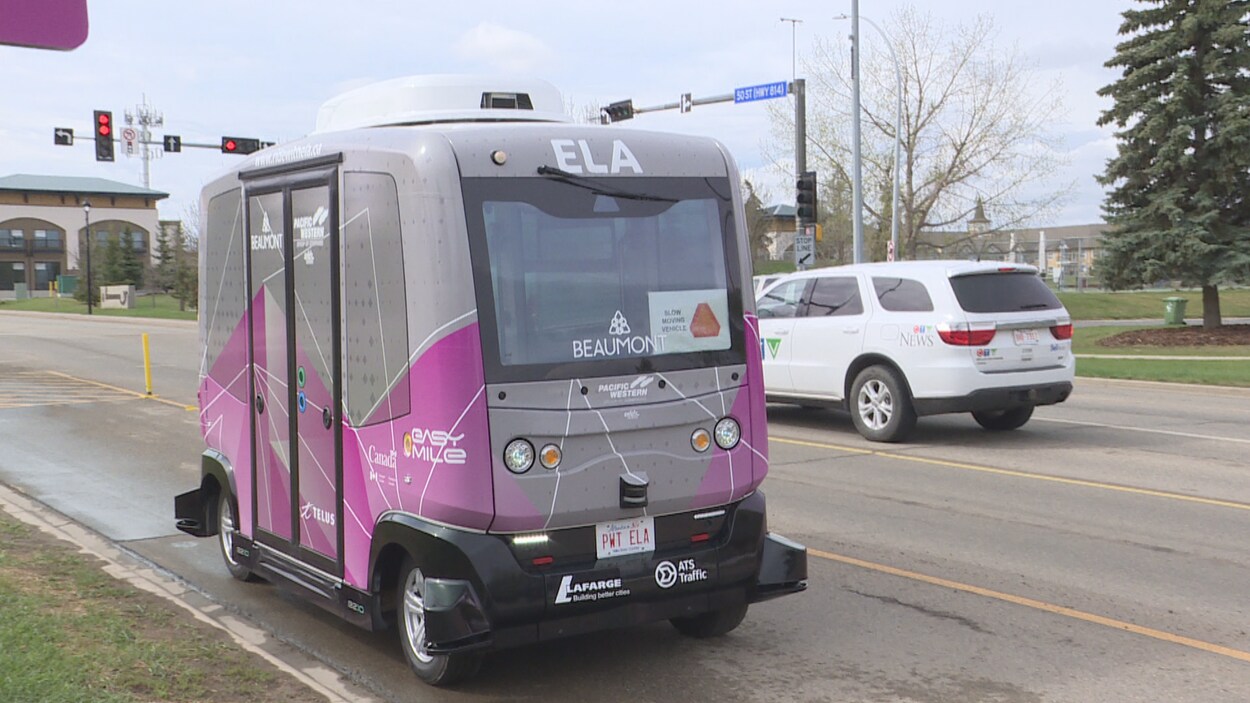 La navette, mauve et grise, roule dans une voie réservée en direction de la caméra. Une voiture et des feux de circulation sont visibles derrière elle.
