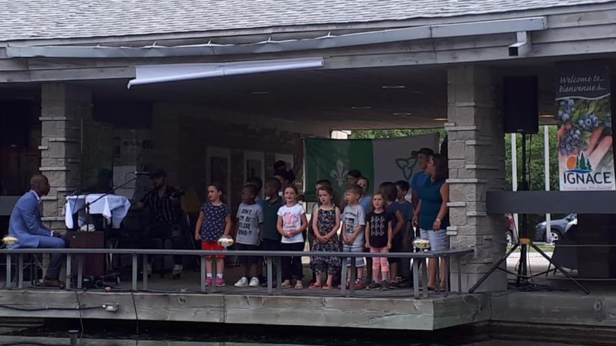 Un groupe d'enfant en train de chanter, accompagné par un pianiste, devant le drapeau franco-ontarien. Un groupe d'enfant en train de chanter, accompagné par un pianiste, devant le drapeau franco-ontarien.