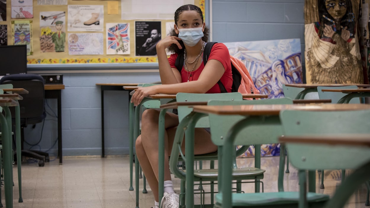 Une élève assise à un pupitre dans une classe vide. Elle porte un masque.