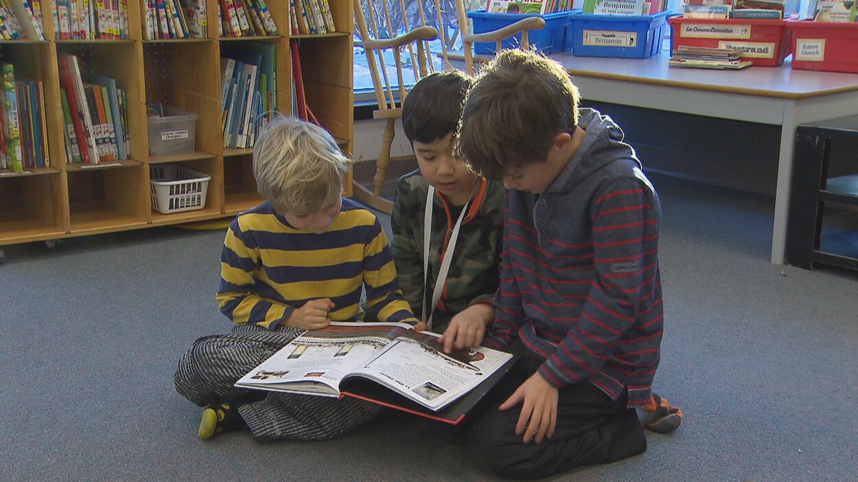 Trois élèves lisent un livre par terre dans une bibliothèque, devant une table et une étagère de livres.