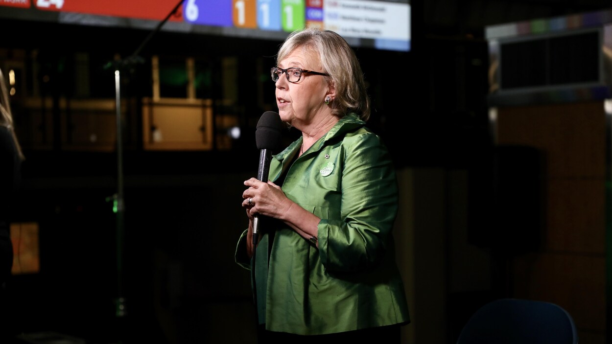 Elizabeth May, micro en main, avec un tableau affichant les résultats électoraux en arrière-plan.