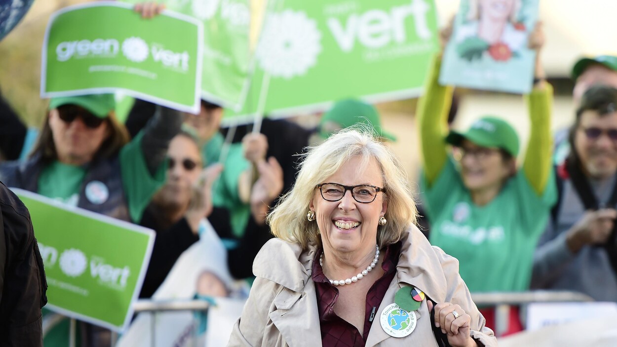 Elizabeth May à son arrivée au débat des chefs. Elle sourit. Derrière elle, des militants du Parti vert brandissent des pancartes.