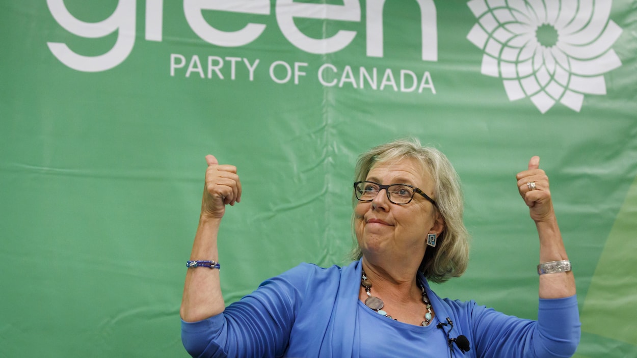 Devant une bannière du Parti vert, Elizabeth May pose, pouces levés vers le ciel.