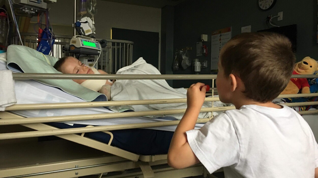 Un jeune garçon est étendu dans un lit d'hôpital alors que son frère jumeau est à ses côtés.