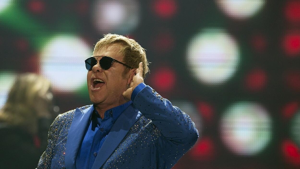 Elton John, sur scène, porte un costume bleu et des lunettes de soleil et écoute la foule chanter. Elton John, sur scène, porte un costume bleu et des lunettes de soleil et écoute la foule chanter.