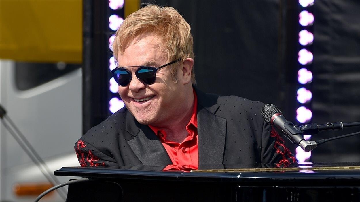 Elton John, assis derrière un piano, sourit en regardant le public. Elton John, assis derrière un piano, sourit en regardant le public.