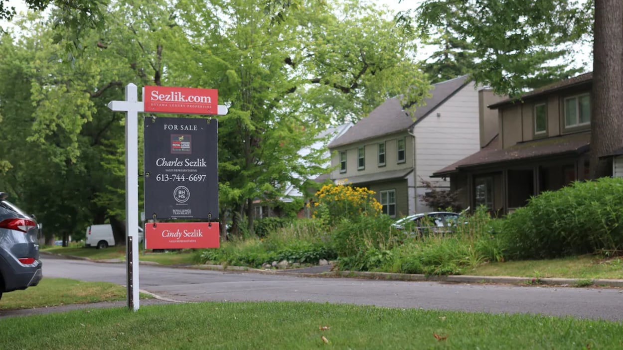Une pancarte indiquant qu'une maison est à vendre sur la pelouse d'une avenue résidentielle. Les coordonnées d'un agent immobilier y figurent.