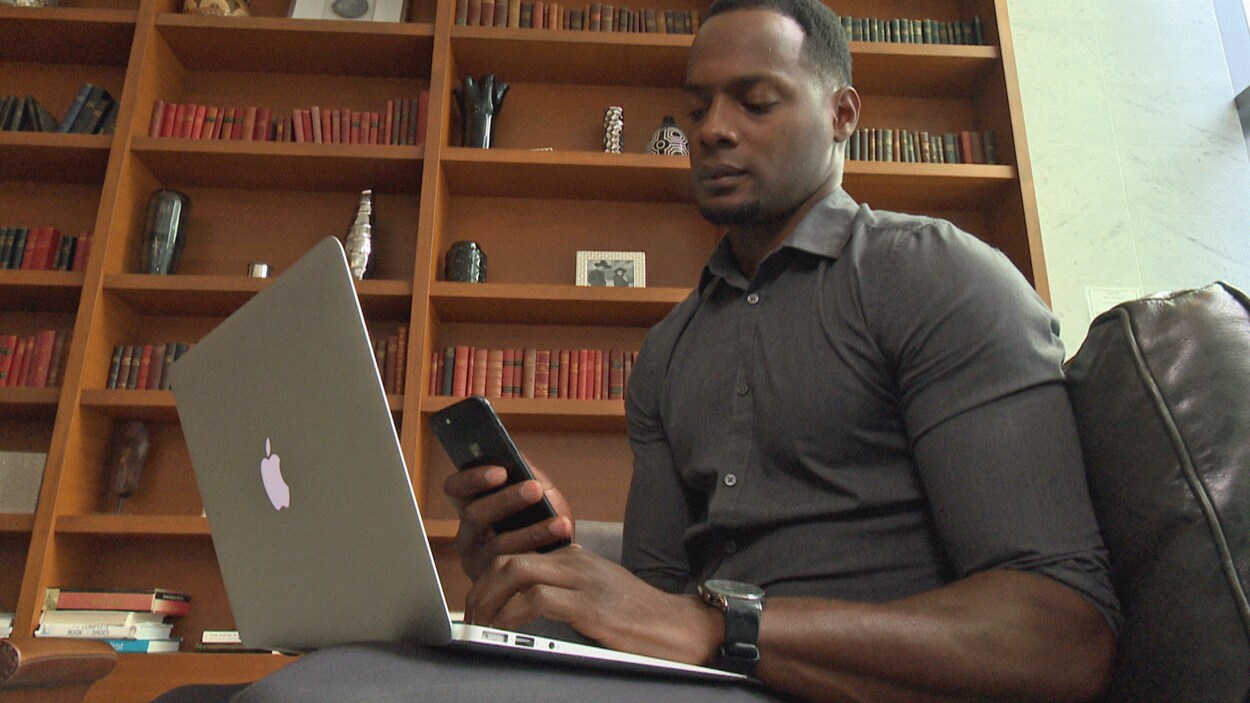 Jermel Pierre assis devant son ordinateur portatif en train d'utiliser son téléphone intelligent.