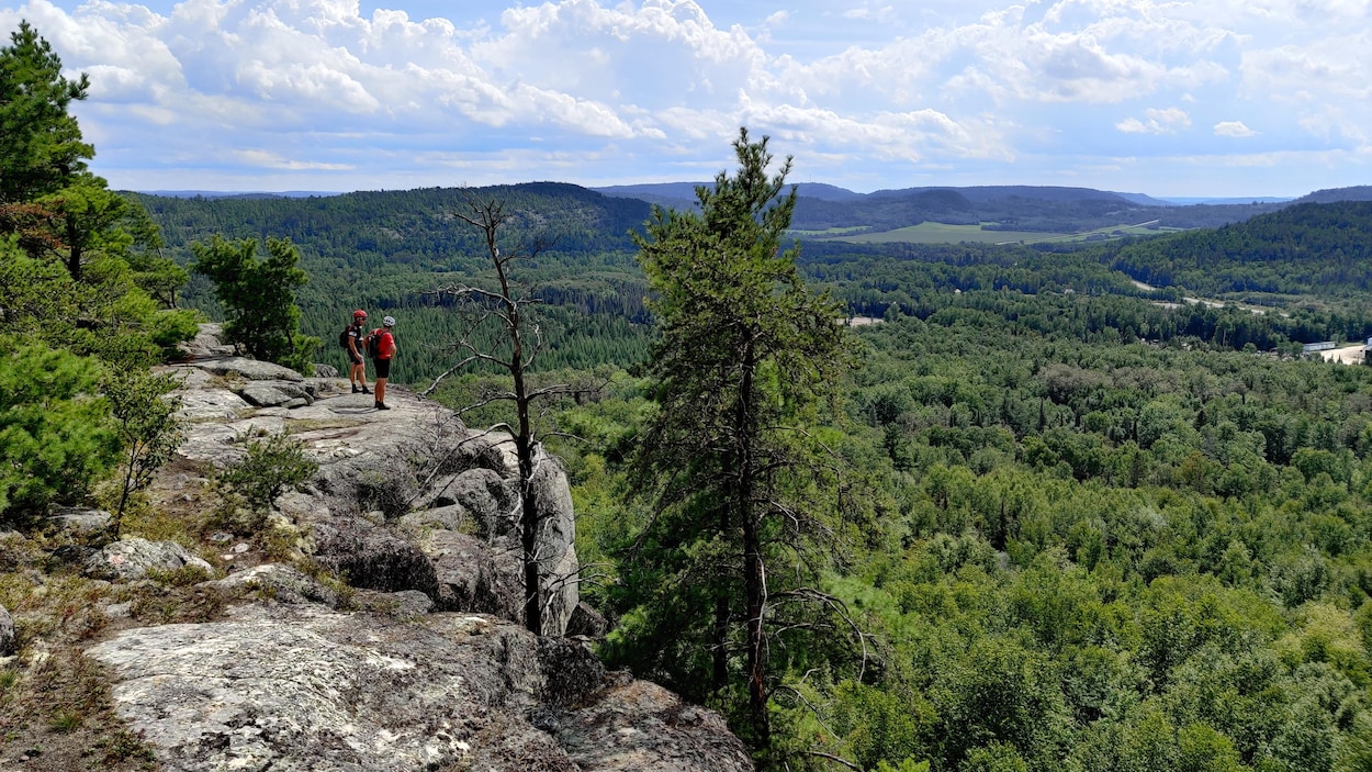 Deux hommes sont debout sur un cap de roche. Ils regardent les arbres à l'horizon et portent un casque de vélo.