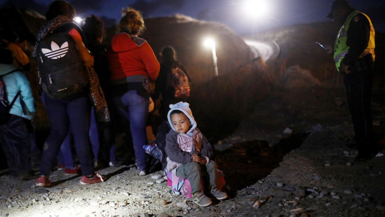 Un jeune enfant est assis sur son sac à dos et fixe le sol pendant que les agents frontaliers américains surveillent un groupe de migrants.
