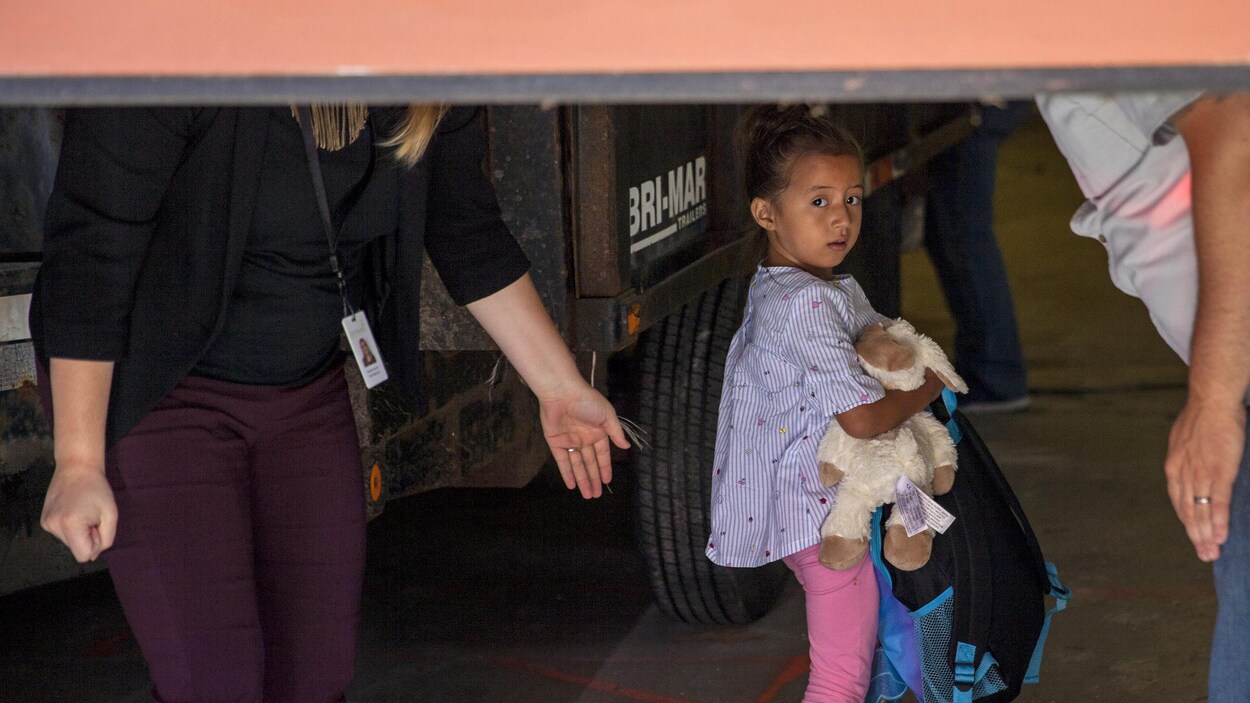 Une petite fille avec un pantalon rose et une chemise tient son doudou et un sac à dos, une femme blanche dont on ne voit pas la tête lui tend la main.