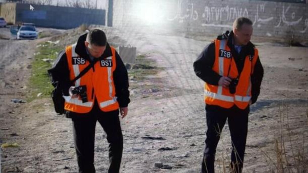 Deux hommes portant des vestes orange sur lesquelles on peut voir les lettres TSB et BST marchent sur un terrain extérieur, appareil photo en main.