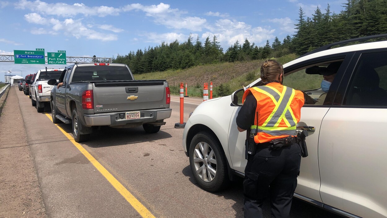 Une femme debout sur l'autoroute, de dos, portant une veste orange et jaune, parle à un automobiliste dans son véhicule.