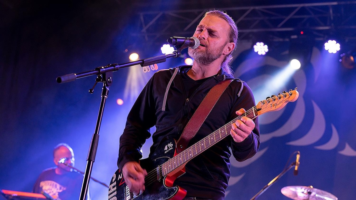 Le chanteur devant le micro, sur scène, qui joue de la guitare.
