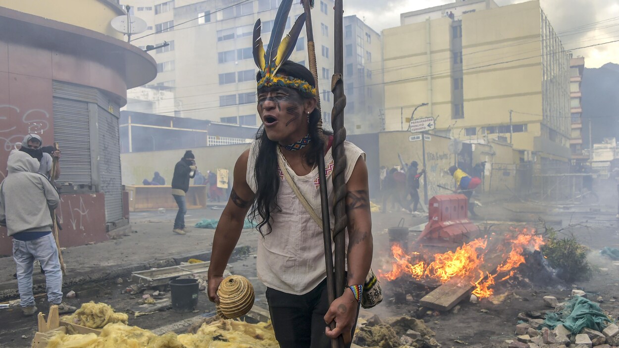 Un homme portant une coiffe de plumes et tenant des lances semble crier, des détritus en flammes derrière lui.