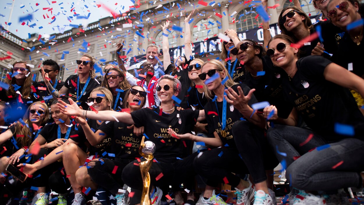 Toutes rassemblées pour une photo de groupe sous les confettis, les joueuses sourient à la caméra. Elles portent toutes un chandail où l'on peut lire « Championnes du monde ».
