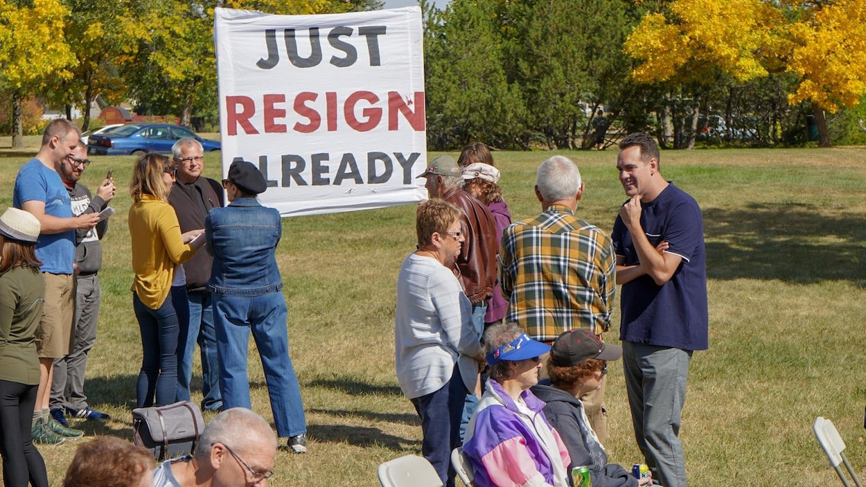 Des personnes dans un parc par une journée ensoleillée. Certaines d'entre elles sont réunies autour d'une banderole où il est noté «Just resign already». Erin Weir se tient à côté, discutant avec une personne en souriant.
