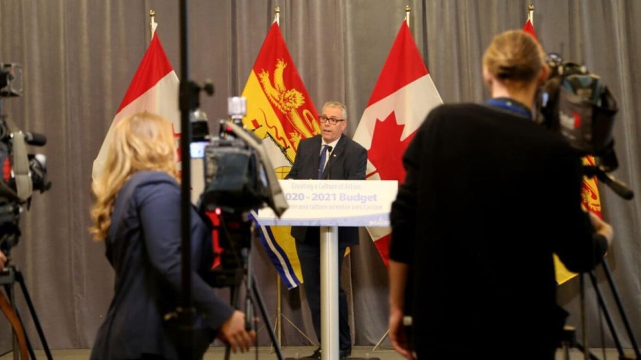 Un homme en habit parle devant plusieurs caméras en conférence de presse.
