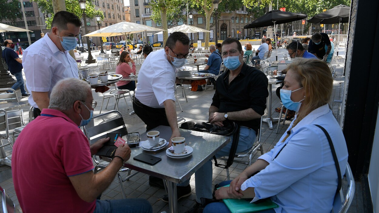 Deux garçons de café débarrassent une table à laquelle sont attablés deux hommes et une femme. Tout le monde est masqué. Les tables sont à bonne distance les unes des autres.