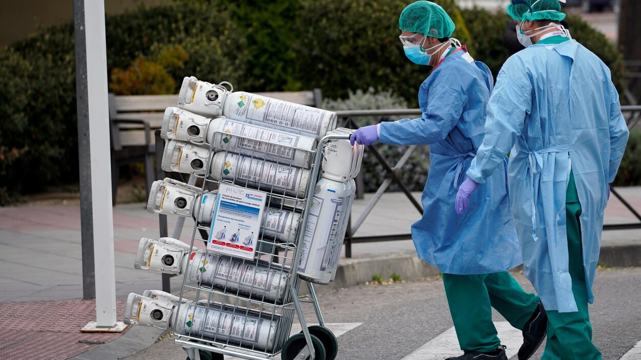 Deux travailleurs de la santé, portant des masques de protection, transportent des bouteilles d'oxygène sur un chariot.