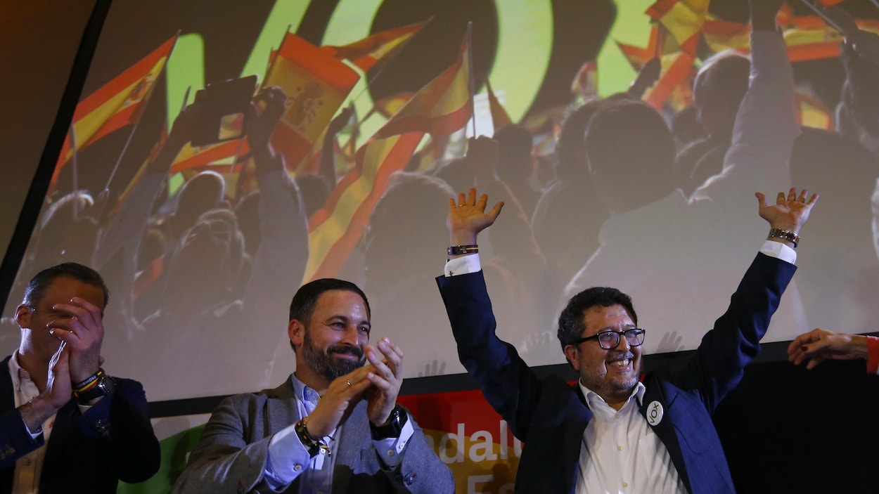 Deux hommes applaudissent alors qu'un autre lève les bras dans les airs devant un écran sur lequel est projeté des images d'un rassemblement politique en Espagne.