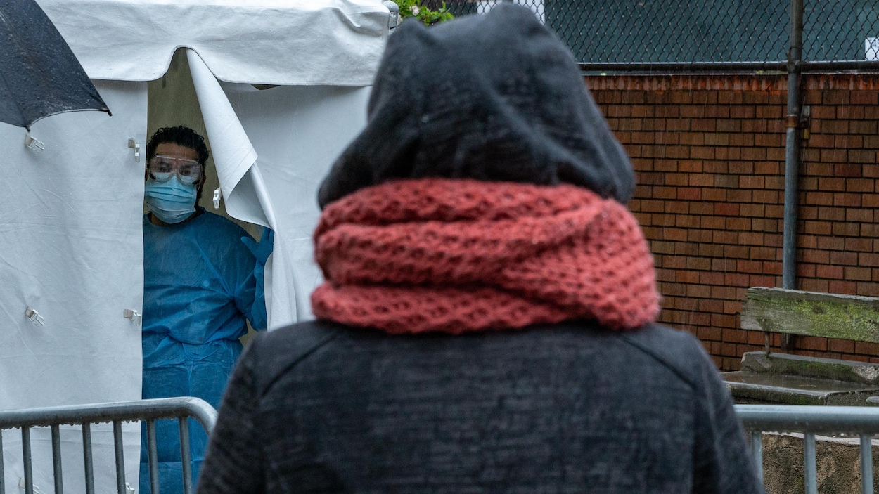 Une femme attend devant une tente où se trouve une personne vêtue d'un masque et d'un habit de protection.