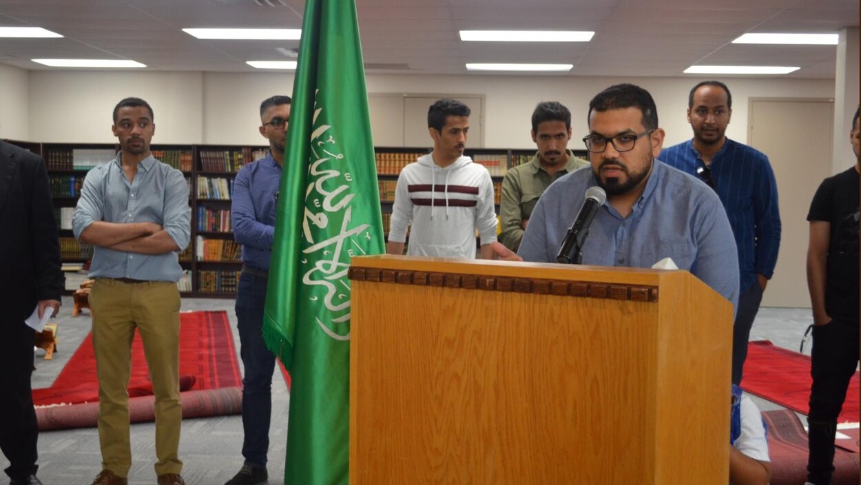 Un homme aux lunettes rectangulaires parle au micro. Au second plan, à sa droite, deux hommes bruns et le drapeau de l'Arabie saoudite.