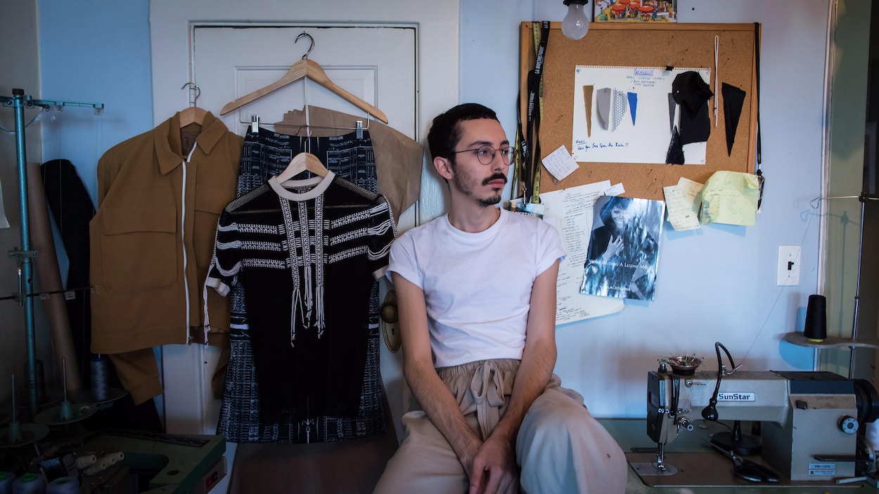 Assis sur un bureau où est installée une machine à coudre, Evan regarde au loin.