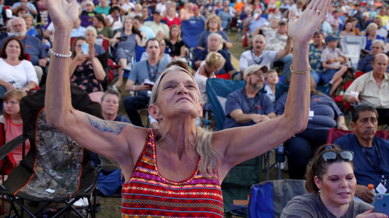 Une foule de personnes sont assises sur des chaises de camping. Une femme est debout, les bras levés vers le ciel.