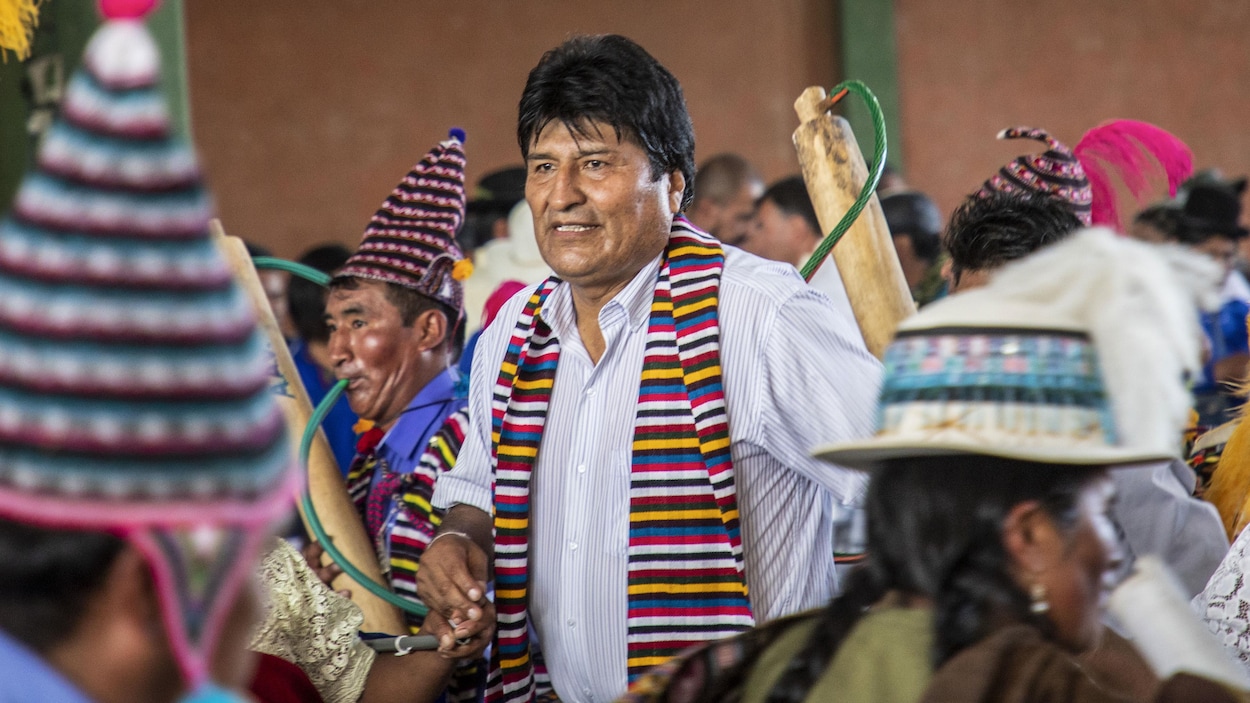 Un homme portant une chemise aux manches retroussées et un foulard multicolore danse avec des Boliviens en tenues traditionnelles.