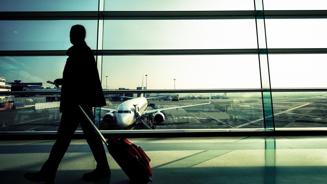 Silhouette d'un homme qui marche en tirant une valise dans un couloir d'aéroport vitré avec vue sur des avions stationnés sur la piste.