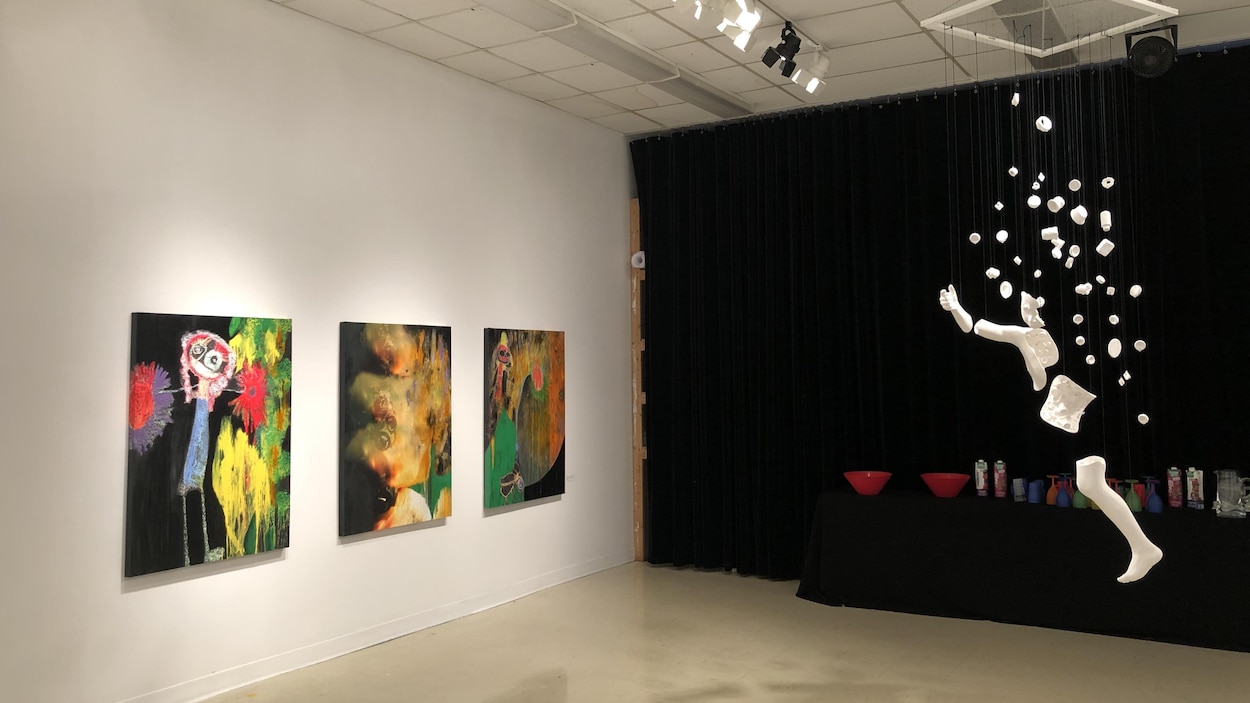 Trois toiles sur un mur et une sculpture qui pend du plafond dans une salle d'exposition.