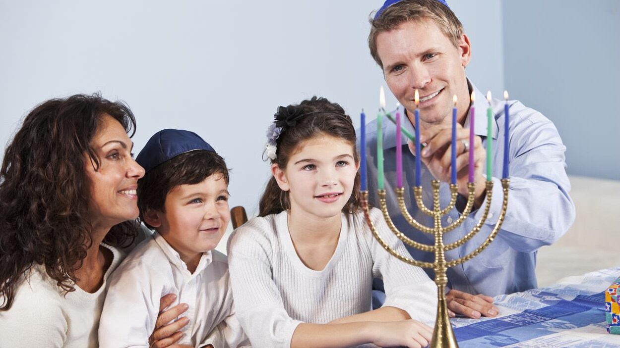Des parents et leurs deux enfants sont attablés. Le père allume les chandelles d'une menora. Des parents et leurs deux enfants sont attablés. Le père allume les chandelles d'une menora.