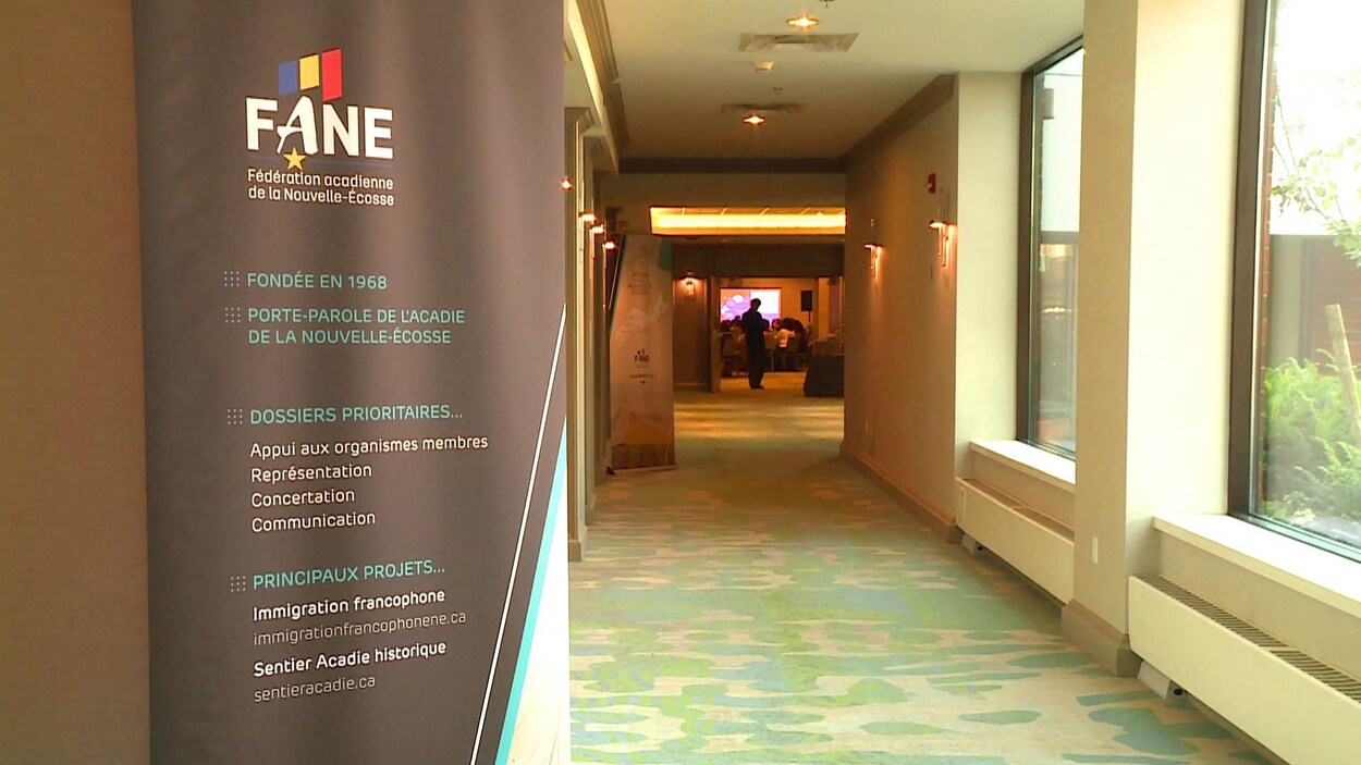 Une bannière de la Fédération acadienne de la Nouvelle-Écosse à l'entrée d'un long corridor qui débouche sur une salle de conférence remplie de gens.