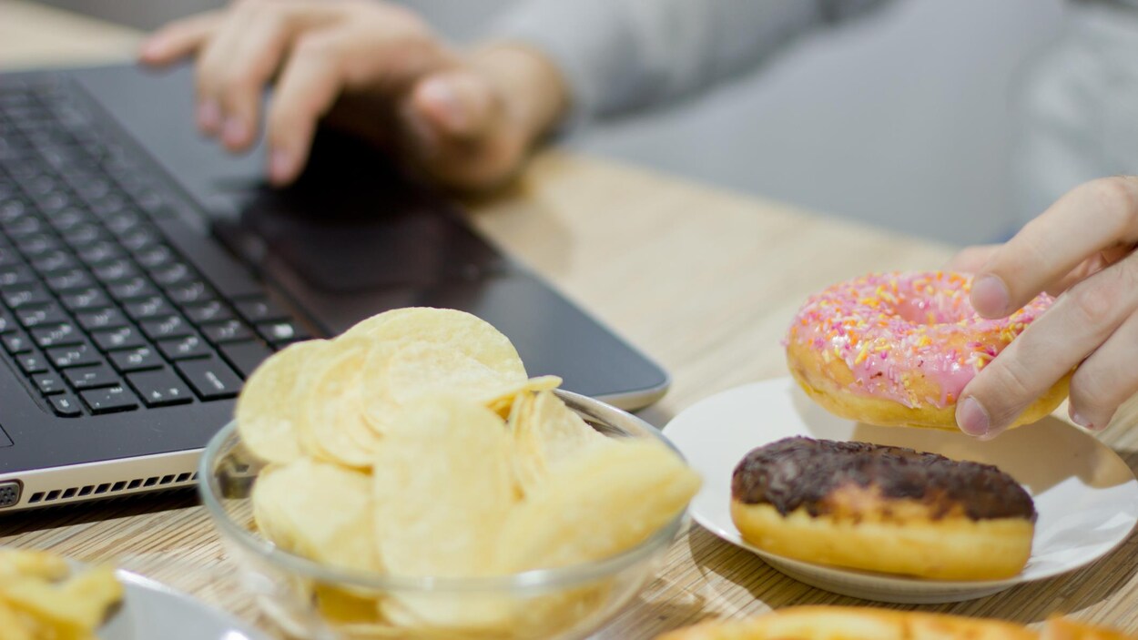 Une personne fait du télétravail en étant entouré de plateaux de chips, de frites, de beignes et de pizzas.