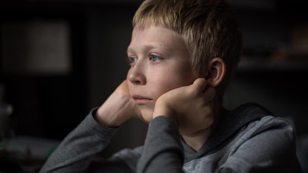 Un enfant blond, sa tête posées dans ses mains, les coudes sur son bureau, regarde au loin, dans une scène du film <i>Faute d'amour</i>.