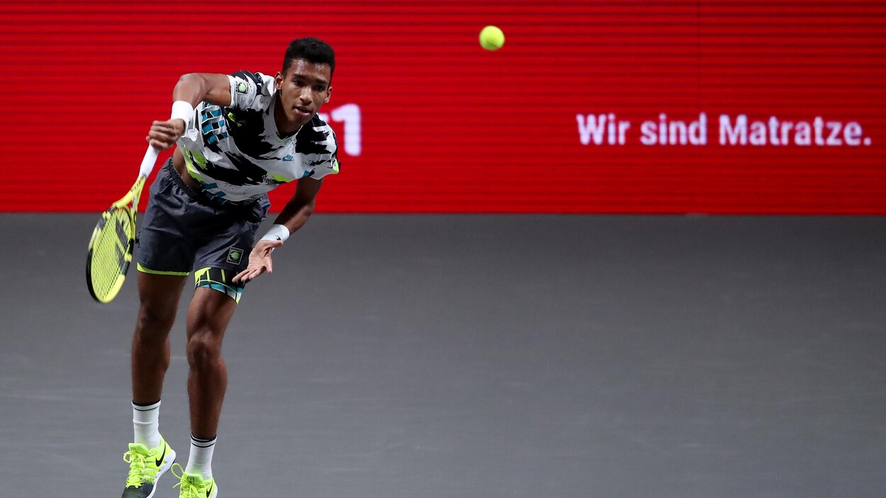 Un joueur de tennis, vêtu de blanc, gris et jaune, frappe un service sur un terrain gris.