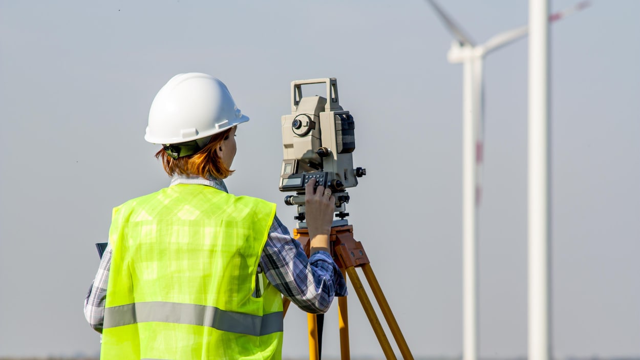 Une arpenteuse-géomètre dans un champ fait des mesures devant une éolienne.