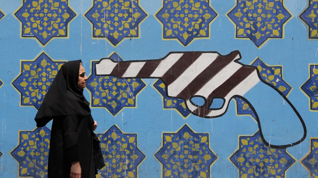Une Iranienne marche devant une murale où l'on voit un pistolet peint aux couleurs du drapeau américain.