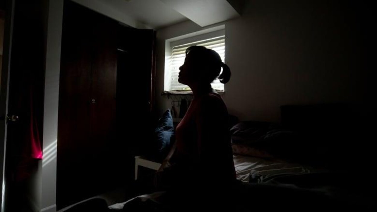 Une femme est assise sur son lit, dans la pénombre du chambre à coucher.