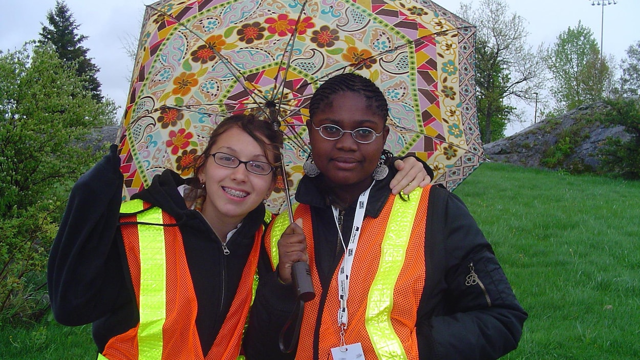 Les deux jeunes participantes portent un parapluie dehors. Elle porte un badge des jeux et un vêtement avec des rubans réfléchissants Les deux jeunes participantes portent un parapluie dehors. Elle porte un badge des jeux et un vêtement avec des rubans réfléchissants