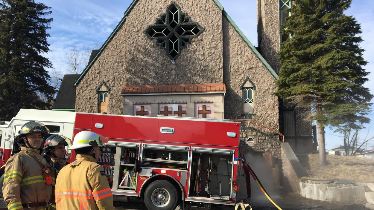 Trois pompiers et un camion de pompier devant l'église Saint-Joachim à Chicoutimi.