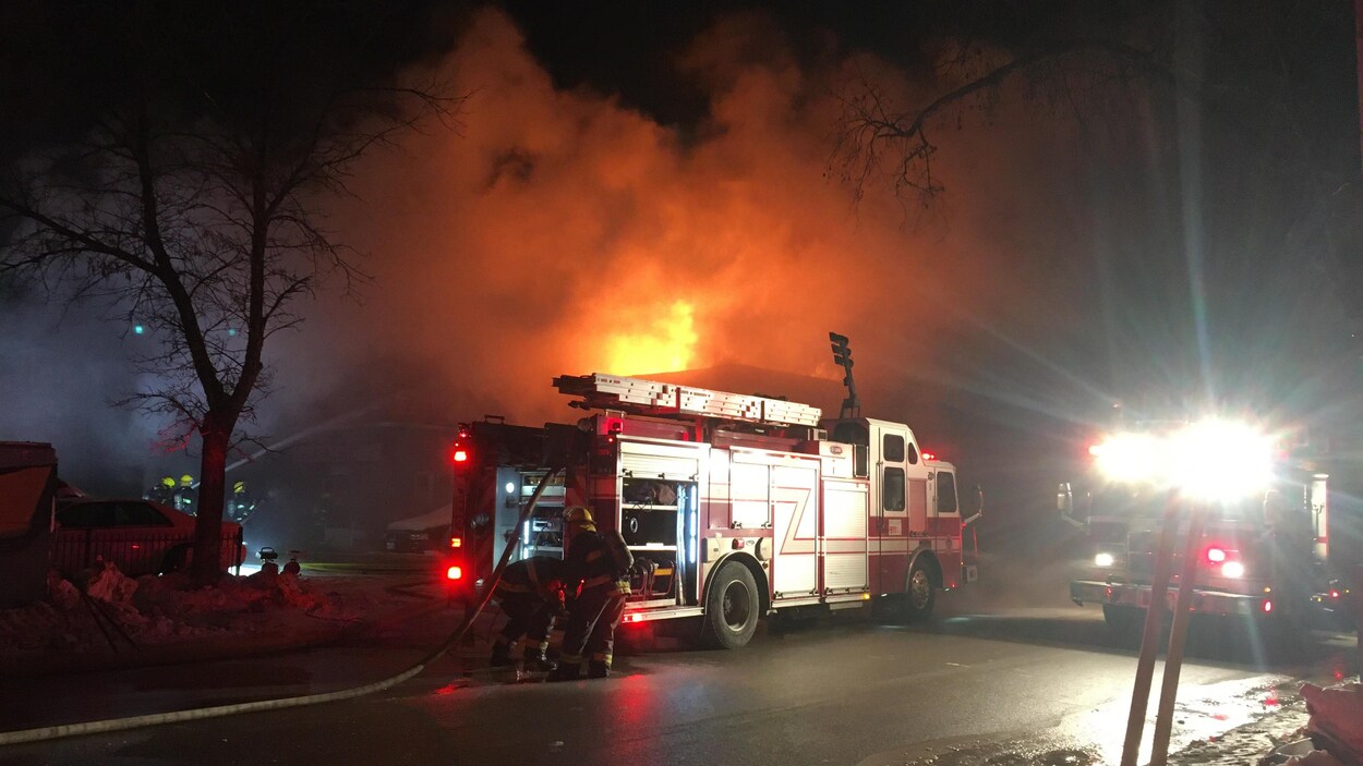 Des pompiers s'affairent autour d'un camion d'incendie dernière lequel on voit des flammes et de la fumée monter dans le ciel.