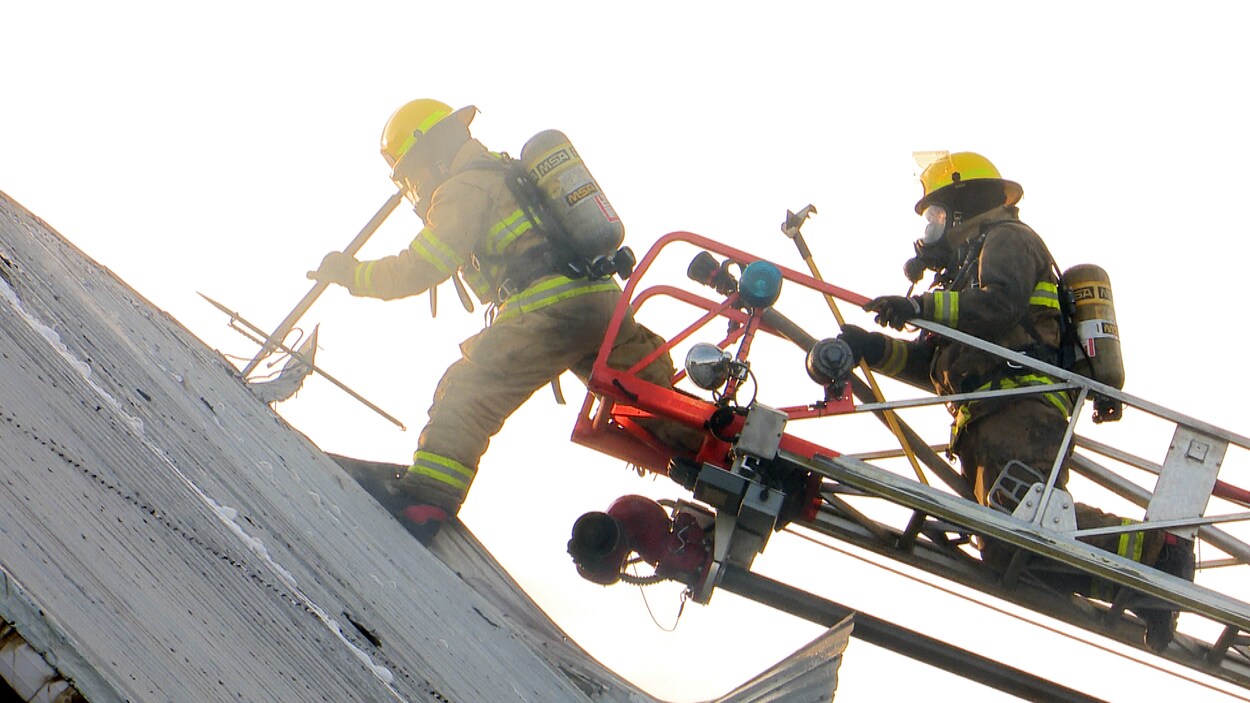 Deux pompiers, au bout de la grande échelle, travaillent près d'un toit.