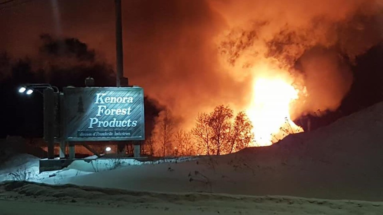 Un brasier illumine le ciel en extérieur devant un panneau de Kenora Forest Products. Un brasier illumine le ciel en extérieur devant un panneau de Kenora Forest Products.