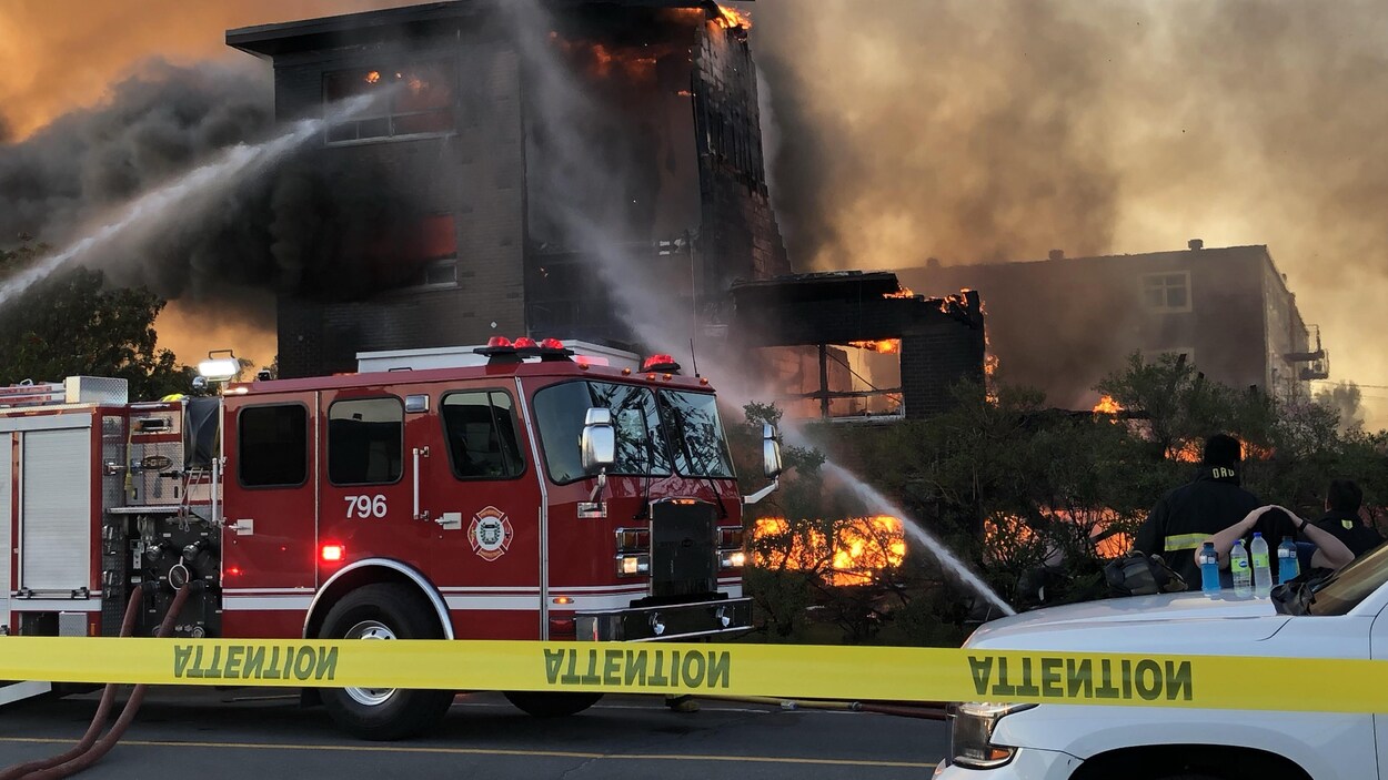 Des flammes jaillissent de l'immeuble à moitié consumé. Au premier plan, un camion de pompier.