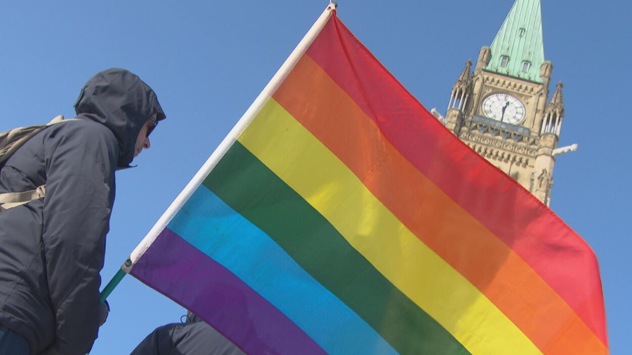Un homme porte le drapeau de la Fierté. Derrière, on peut apercevoir la tour centrale du Parlement.