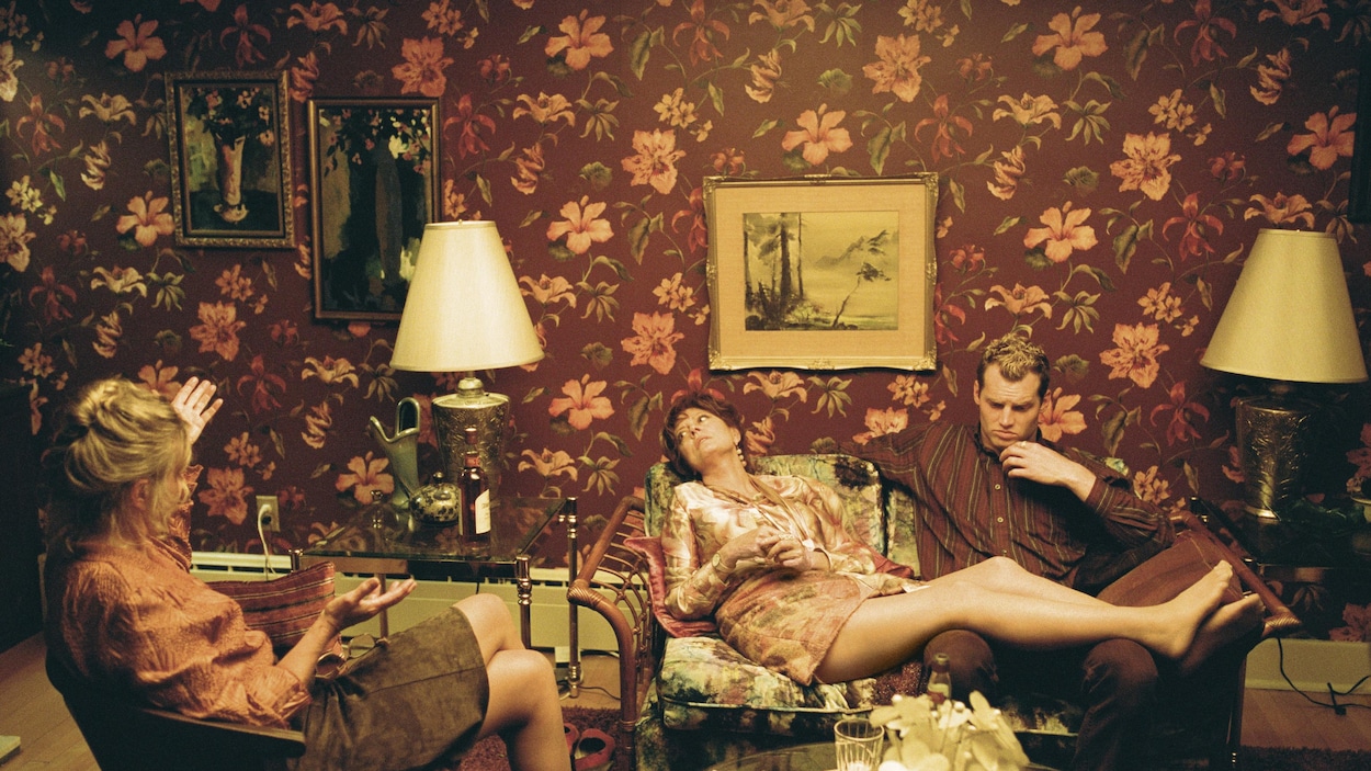 Deux femmes et un homme sont assis sur un sofa dans une pièce couverte de tapisserie fleurie.