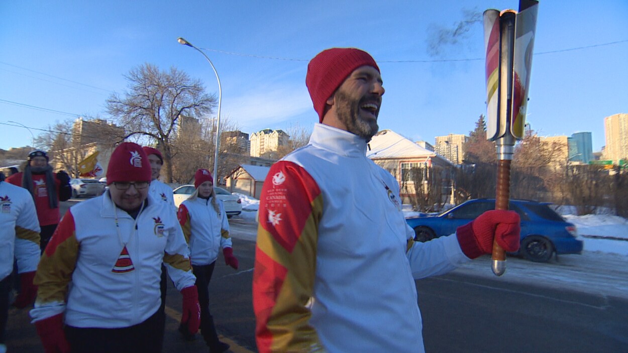 Don Iveson porte un flambeau, dehors. Il semble être en train de rire. Une petite foule habillée aux couleurs des Jeux du Canada le suit.