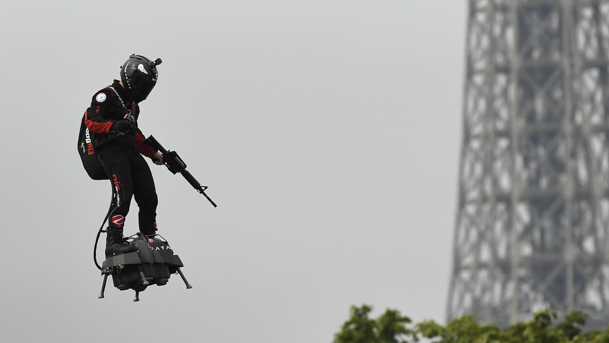 Un homme en combinaison et portant un casque vole à l'aide d'une petite plateforme noire accrochée à ses pieds. L'homme tient un fusil d'assaut dans sa main gauche. En arrière plan, une partie de la tour Eiffel est visible.